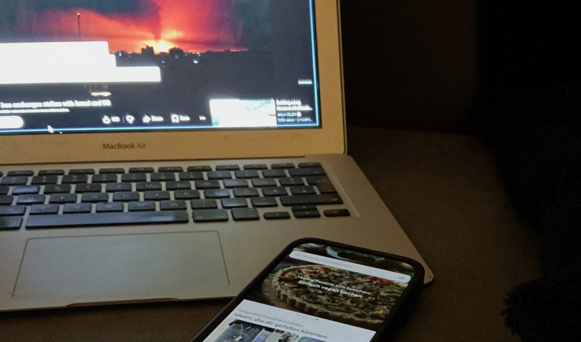 A smartphone lies on a dark table in front of an open laptop with a red-glowing news image on the screen.